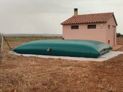 Depósito flexible de agua instalado sobre una base preparada junto a una caseta con cubierta de teja, en una finca agrícola de secano